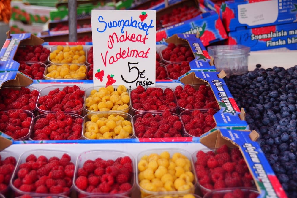 Red and yellow Finnish raspberries displayed beside dark blueberries at a colourful Helsinki market stall, highlighting the fresh summer berries sold by the container in Finland.
