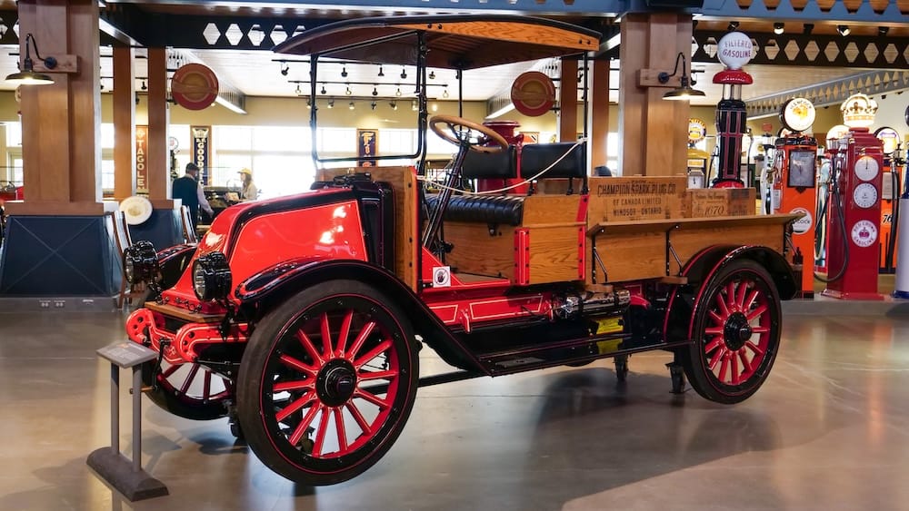 Restored red vintage truck with a wooden cargo bed displayed inside Gasoline Alley Museum at Heritage Park in Calgary, Alberta, Canada, with historic gasoline pumps visible in the background.