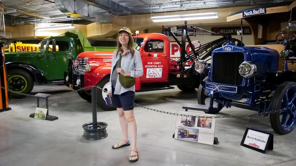 Audrey Bergner standing beside restored vintage trucks inside Gasoline Alley Museum at Heritage Park in Calgary, Alberta, Canada, with colourful historic commercial vehicles displayed behind her.