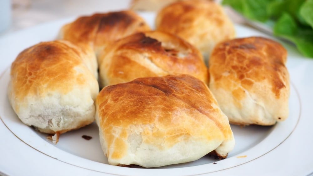 Close-up of golden baked stuffed pastries arranged on a plate during a shared meal in Karakol, Kyrgyzstan, with browned tops and soft bread-like crusts surrounding an unseen savoury filling.

