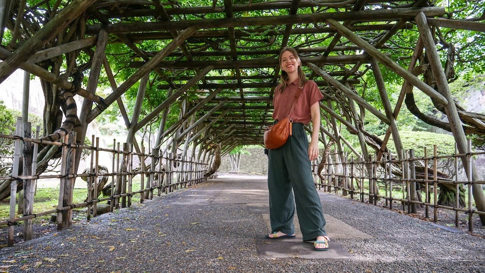 Audrey Bergner bajo una pérgola de madera cubierta de hojas en el parque Goryokaku, Hakodate, Hokkaido, Japón Audrey Bergner bajo una larga pérgola de madera cubierta de hojas dentro del parque Goryokaku, en Hakodate, Hokkaido, Japón, con senderos de grava y fortificaciones históricas de piedra cerca.