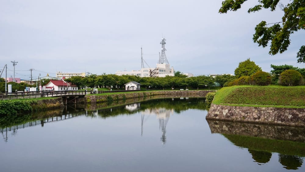 Foso quieto con pared de piedra y puente de madera, Parque Goryokaku, Hakodate, Hokkaido, Japón Un foso tranquilo en el Parque Goryokaku en Hakodate, Hokkaido, Japón, refleja una muralla de pasto con paredes de piedra y un puente de madera, con una cabañita, un edificio de techo rojo y los edificios de la ciudad de fondo bajo un cielo gris.