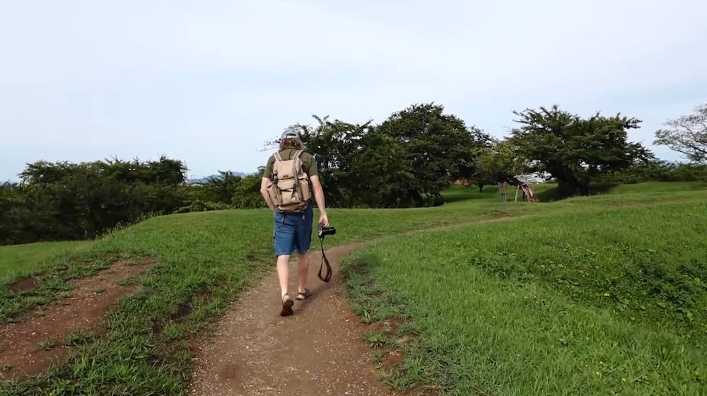 Samuel Jeffery walks along a narrow dirt path through the grassy grounds of Goryokaku Park in Hakodate, Hokkaido, Japan, carrying a camera and backpack with trees spread across the fort landscape.