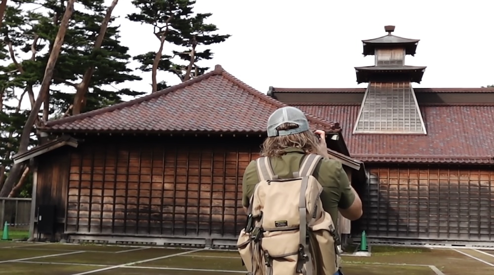 Samuel Jeffery Photographing Historic Wooden Architecture at Goryokaku Park in Hakodate, Hokkaido, Japan Samuel Jeffery photographing a traditional Japanese wooden building at Goryokaku Park in Hakodate, Hokkaido, Japan, wearing a baseball cap and beige backpack, with tiled roofs, a raised tower and pine trees behind him.
