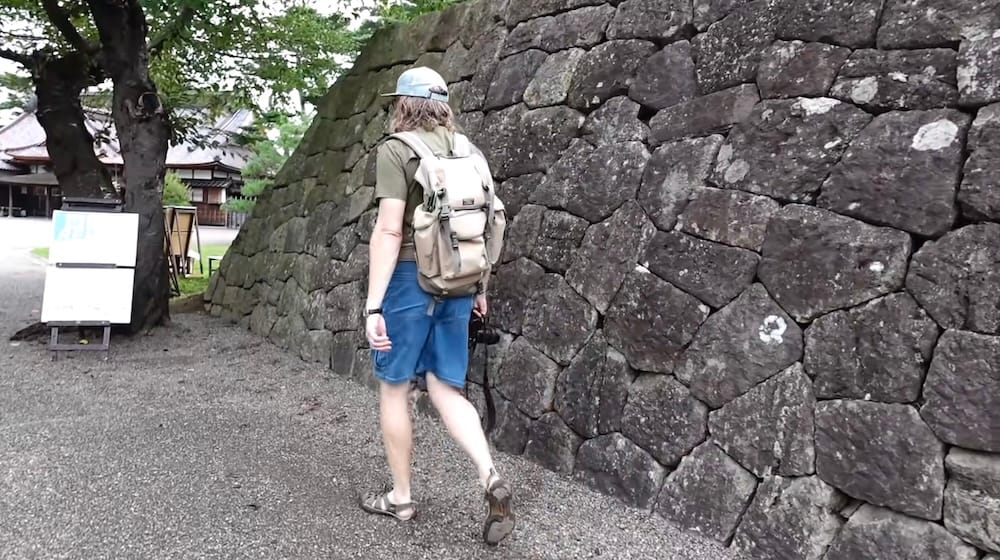 Samuel Jeffery, seen from behind in a cap and backpack, walks along a gravel path beside a large stone wall in Goryokaku Park in Hakodate, Hokkaido, Japan, with a tiled-roof building and trees beyond.