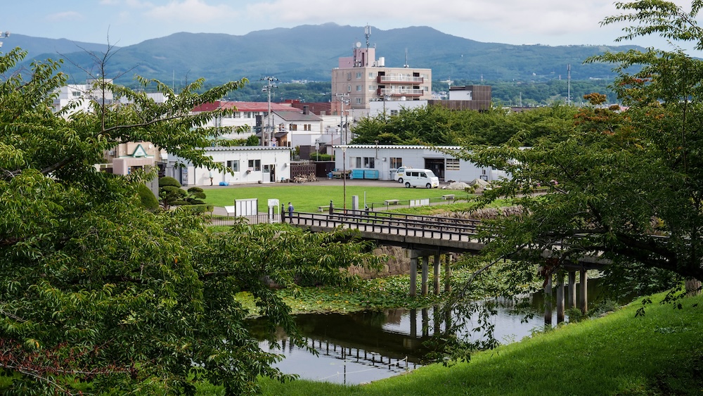Foso y puente del Parque Goryokaku con edificios de Hakodate y montañas en Hokkaido, Japón Vista desde el Parque Goryokaku en Hakodate, Hokkaido, Japón, con un puente sobre el foso, árboles y césped verdes, edificios cercanos y montañas elevándose detrás del barrio.
