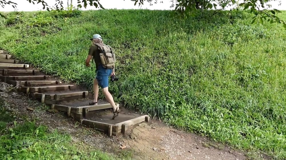 Samuel Jeffery sube escalones de madera entre los espacios verdes de Goryokaku Park, en Hakodate, Hokkaido, Japón, con una cámara y una mochila junto a un terraplén cubierto de césped.