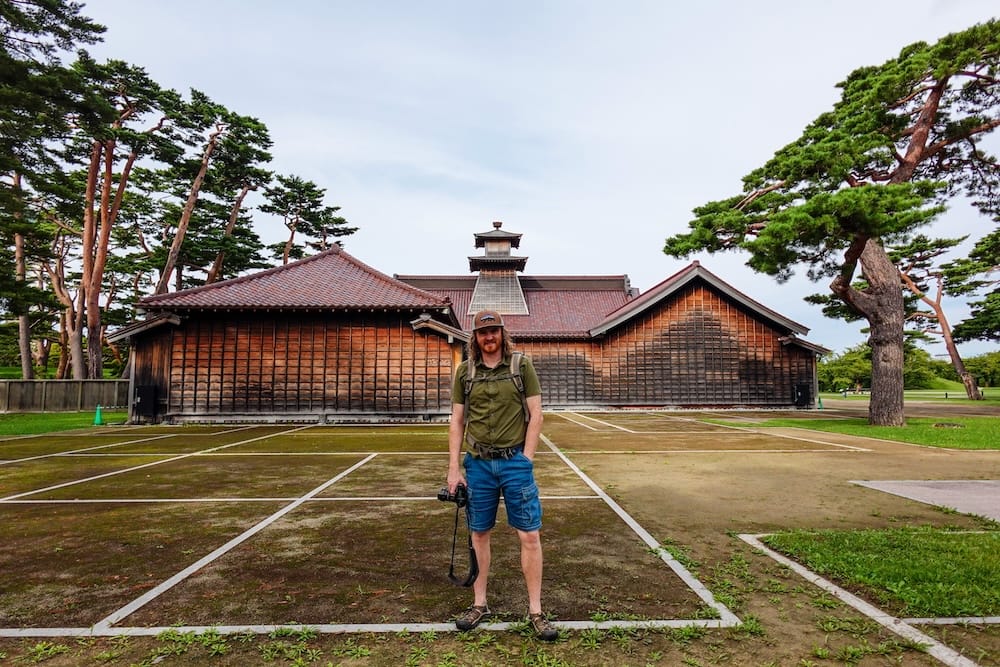 Samuel Jeffery está frente a un edificio histórico de madera en Goryokaku Park, Hakodate, Hokkaido, Japón, con espacios abiertos y altos pinos que muestran una zona más tranquila y con menos gente.