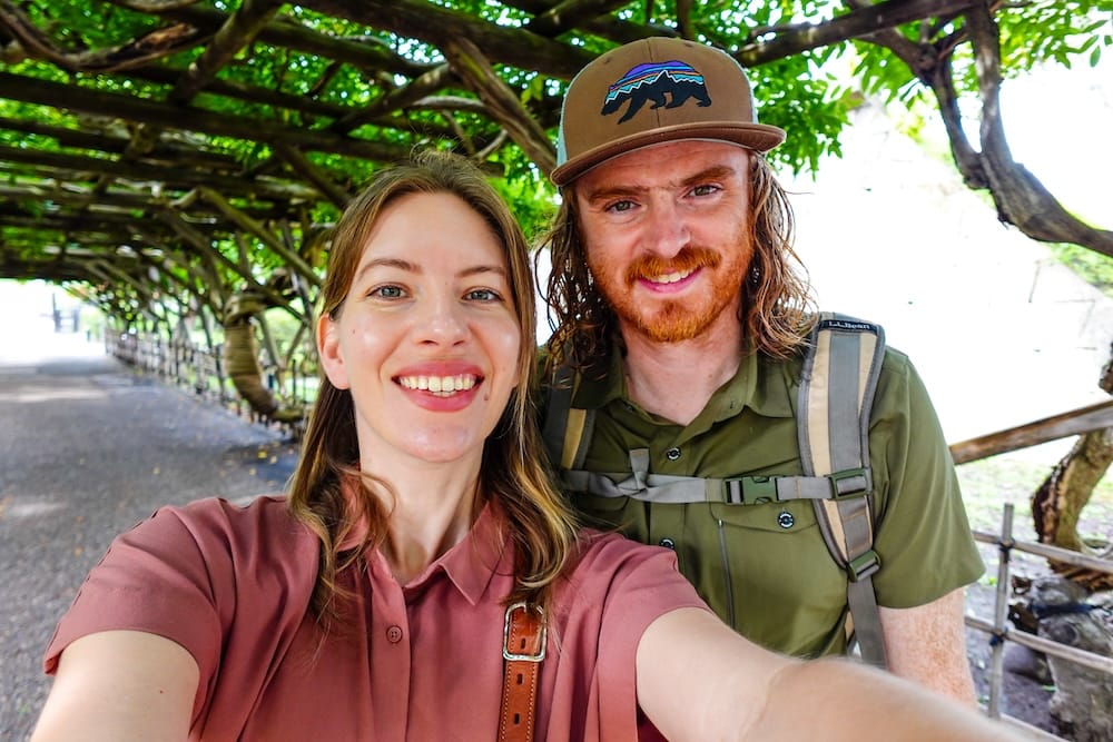 Samuel Jeffery and Audrey Bergner smile for a selfie under a green wisteria trellis in Goryokaku Park in Hakodate, Hokkaido, Japan, with a gravel path and wooden fence visible behind them.