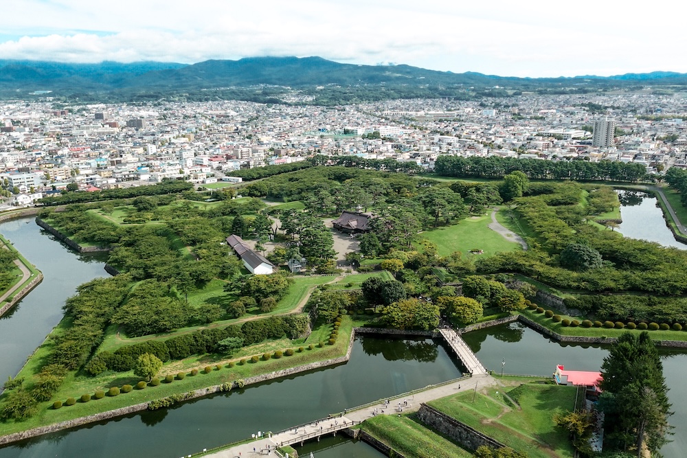 Goryokaku Star Fort From Observation Tower, Hakodate, Hokkaido, Japan – Moats, Earthworks and Historic Fortifications Elevated view from Goryokaku Tower in Hakodate, Hokkaido, Japan, showing the star-shaped fortress's angular stone walls, green defensive earthworks, water-filled moats, bridges and reconstructed Magistrate's Office surrounded by the city.
