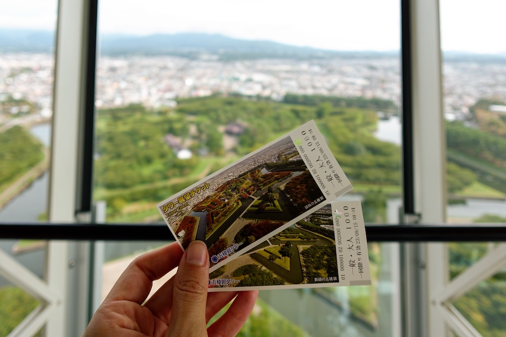 Goryokaku Tower Admission Tickets with Views of the Historic Star-Shaped Fort in Hakodate, Hokkaido, Japan Audrey Bergner holding two Goryokaku Tower admission tickets in front of the observation deck windows in Hakodate, Hokkaido, Japan, with the green star-shaped fort, surrounding moat and city blurred in the background.