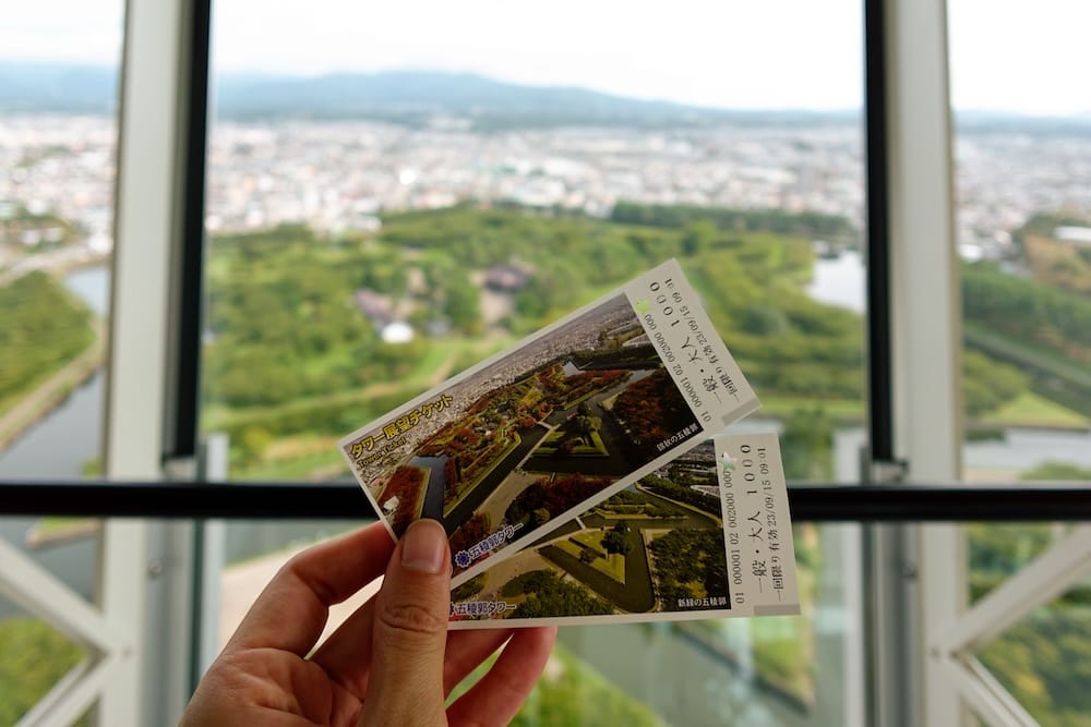 Entradas de Goryokaku Tower con el fuerte en estrella visible abajo, en Hakodate, Hokkaido, Japón Dos entradas de Goryokaku Tower sostenidas dentro de la plataforma de observación en Hakodate, Hokkaido, Japón, con el fuerte en forma de estrella, el foso y la ciudad desenfocados detrás de los ventanales.