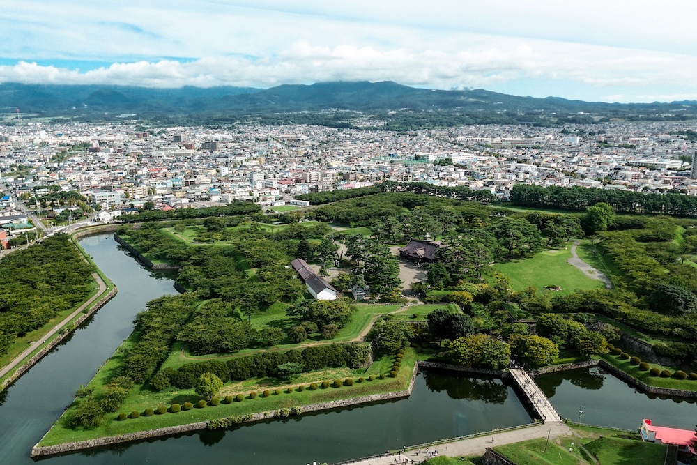 Elevated view from Goryokaku Tower in Hakodate, Hokkaido, Japan, showing the historic star-shaped fort's green earthworks, water-filled moats, walking paths and bridge, with city neighbourhoods and mountains beyond.
