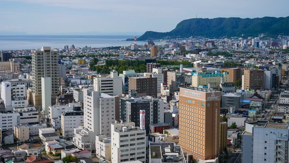 Goryokaku Tower view toward Mt. Hakodate, city centre, coastline and harbour in Hokkaido, Japan View from Goryokaku Tower in Hakodate, Hokkaido, Japan, across dense city buildings toward Mt. Hakodate, the coastline and harbour, with the mountain rising prominently beyond the urban centre.