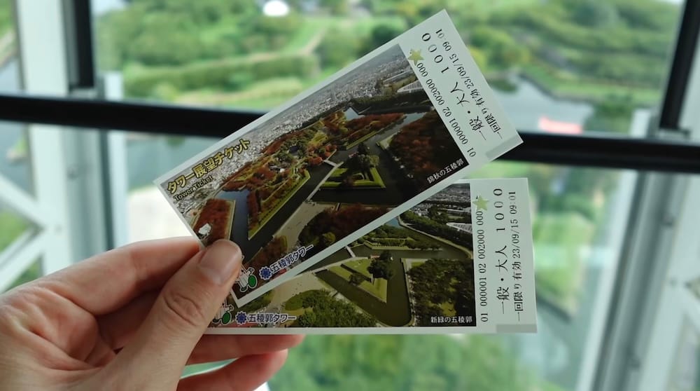 A hand holds two Goryokaku Tower admission tickets inside the observation deck in Hakodate, Hokkaido, Japan, with the green star-shaped Goryokaku grounds visible through the windows behind.
