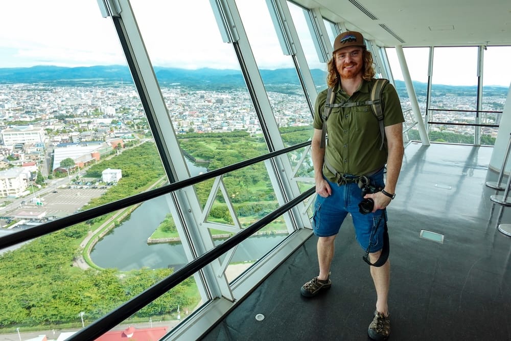 Samuel Jeffery standing with a camera inside the Goryokaku Tower observation deck in Hakodate, Hokkaido, Japan, with the fort moat, green parkland, cityscape and mountains visible through the windows.
