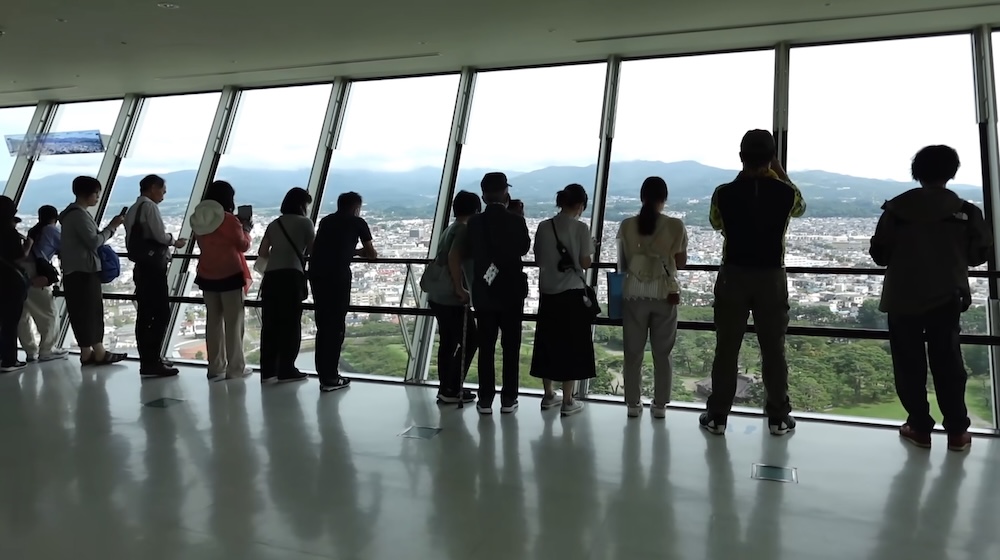 Visitors Enjoying Panoramic City and Mountain Views from Goryokaku Tower Observation Deck in Hakodate, Japan Visitors standing beside large panoramic windows on the Goryokaku Tower observation deck in Hakodate, Hokkaido, Japan, looking out over city buildings, green parkland and distant mountains beneath a cloudy sky.