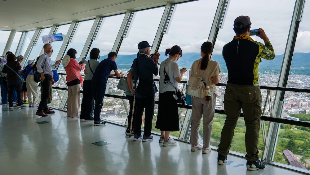 Goryokaku Tower Observation Deck in Hakodate Hokkaido Japan With Visitors Photographing Panoramic City Views Visitors enjoying the views from Goryokaku Tower's indoor observation deck in Hakodate, Hokkaido, Japan, photographing the city through tall angled glass windows, with buildings, green parkland and distant mountains visible.