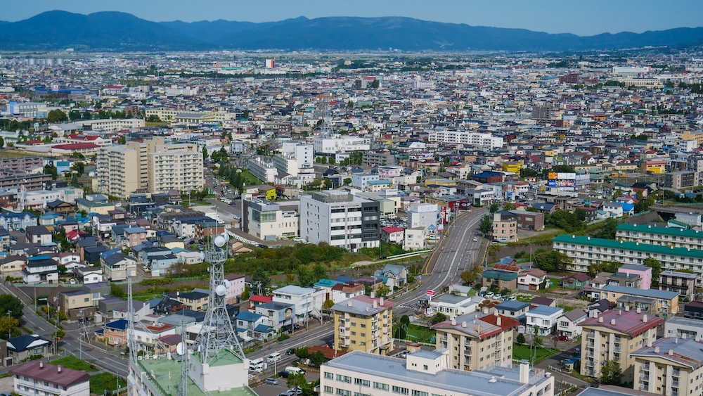 Elevated view from Goryokaku Tower in Hakodate, Hokkaido, Japan, showing densely packed residential neighbourhoods, apartment buildings, intersecting roads, a communications mast and distant mountains.