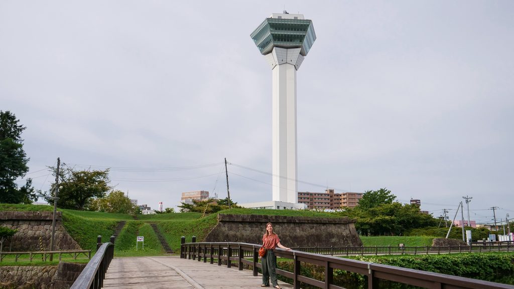Audrey Bergner standing on a wooden bridge in Goryokaku Park in Hakodate, Hokkaido, Japan, with the fortifications, grassy embankments and tall Goryokaku Tower rising behind her under an overcast sky.