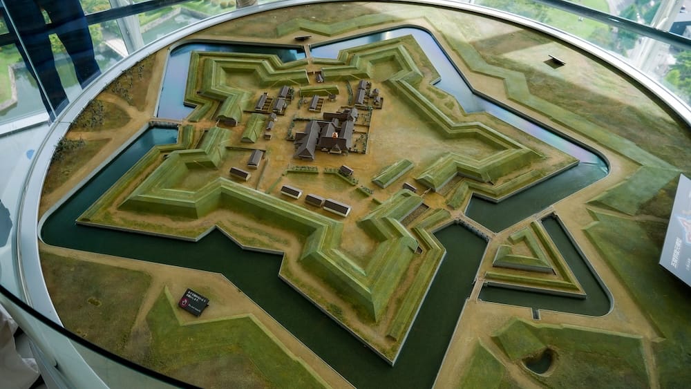 Una maqueta a escala del fuerte en forma de estrella Goryokaku en Hakodate, Hokkaido, Japón, vista desde arriba a través de una baranda de vidrio curva, mostrando el foso de cinco puntas, las murallas verdes y un grupo de edificios en el centro.