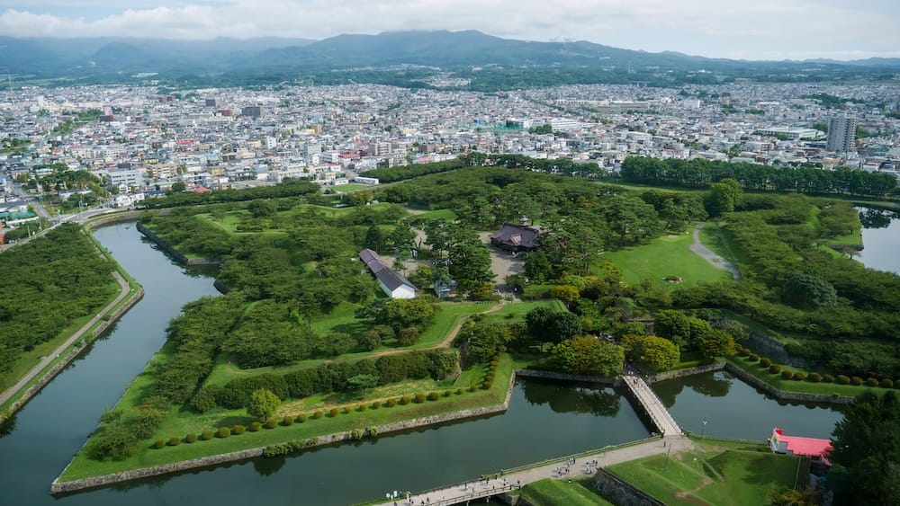 Goryokaku Tower view over the star fort, moat, Hakodate city and distant mountains in Hokkaido, Japan View from Goryokaku Tower in Hakodate, Hokkaido, Japan, looking down over the green star-fort grounds, surrounding moat and bridges, with dense city neighbourhoods and mountains beyond.