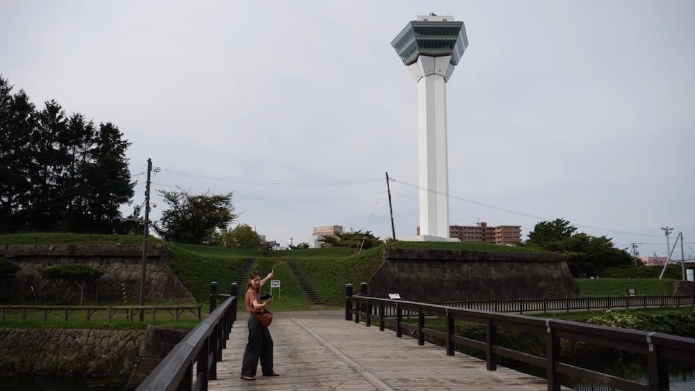 Torre Goryokaku vista desde un puente del foso con paredes de piedra y murallas, Parque Goryokaku, Hakodate, Hokkaido, Japón – Audrey Bergner Audrey Bergner está parada en un puente de madera en el Parque Goryokaku en Hakodate, Hokkaido, Japón, señalando la alta Torre Goryokaku blanca detrás de murallas de pasto, paredes de piedra y un foso bajo un cielo nublado.