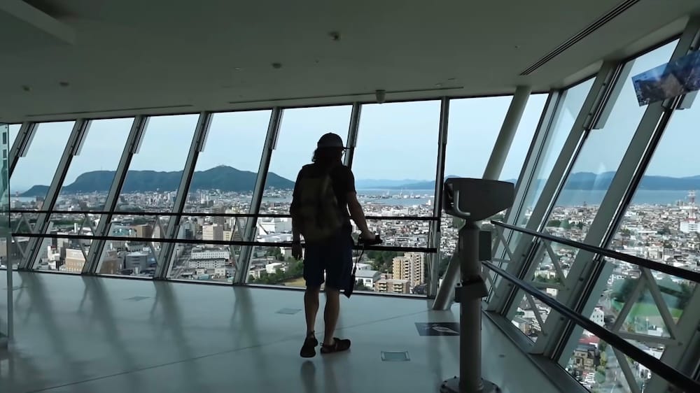 Samuel Jeffery está parado frente a los ventanales de vidrio en la plataforma de observación de la Torre Goryokaku en Hakodate, Hokkaido, Japón, mirando por encima de la ciudad y el puerto hacia una montaña en el horizonte, con un visor que funciona con monedas en primer plano.