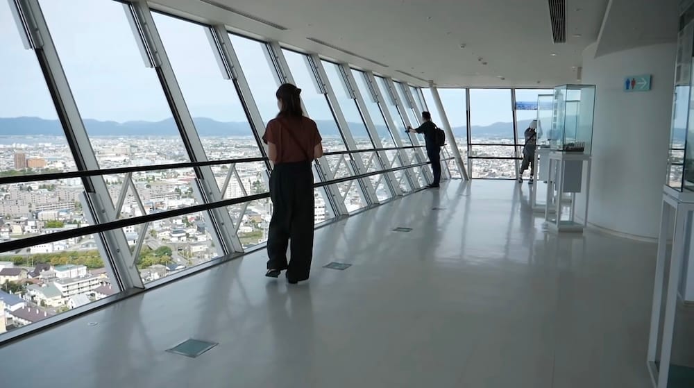 Goryokaku Tower observation deck city view, Hakodate, Hokkaido, Japan – Audrey Bergner Audrey Bergner looks out over Hakodate, Hokkaido, Japan, from the slanted-glass observation deck of Goryokaku Tower, with a sprawling cityscape and distant mountains visible through the windows.