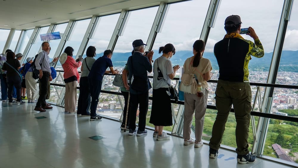 Visitors photograph the view from the slanted-glass observation deck of Goryokaku Tower in Hakodate, Hokkaido, Japan, looking out over the city, green parkland below and distant mountains under a cloudy sky.