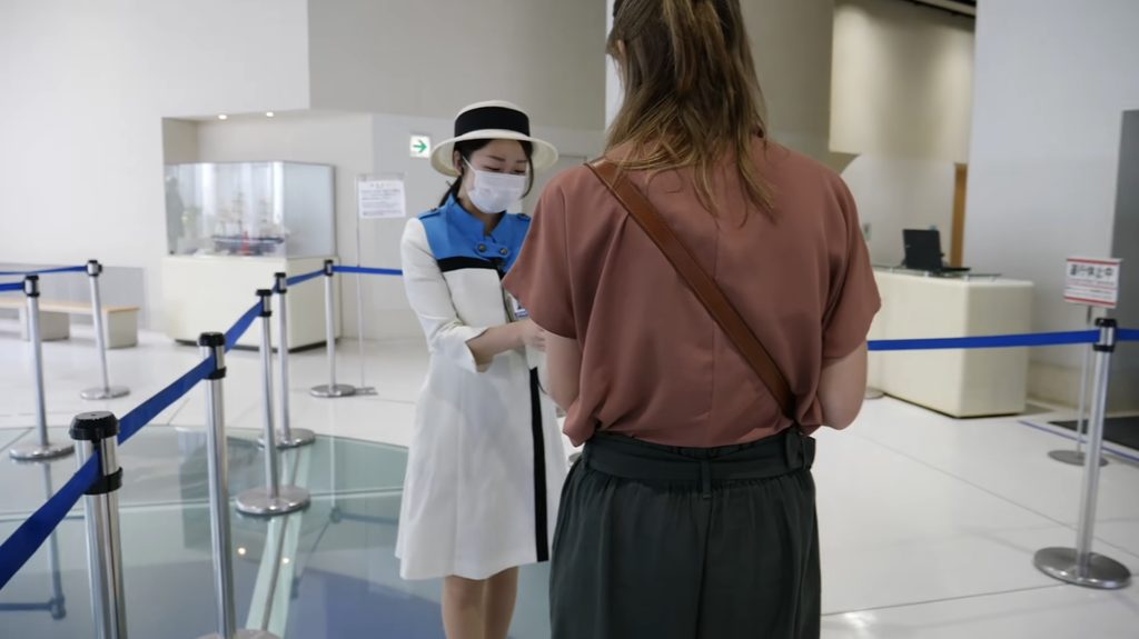 Ticket check at Goryokaku Tower lobby, Hakodate, Hokkaido, Japan – Audrey Bergner A uniformed staff member checks tickets for Audrey Bergner in the lobby of Goryokaku Tower in Hakodate, Hokkaido, Japan, with a roped queue line and a model ship display in the background.