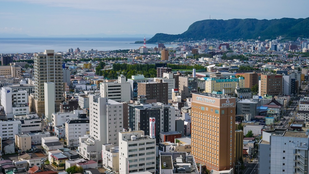 Panorama desde la Torre Goryokaku sobre Hakodate, Hokkaido, Japón, con puerto, ciudad y Monte Hakodate Vista desde la Torre Goryokaku sobre Hakodate, Hokkaido, Japón, con barrios urbanos densos, el puerto, la costa y el Monte Hakodate elevándose detrás del perfil de la ciudad.