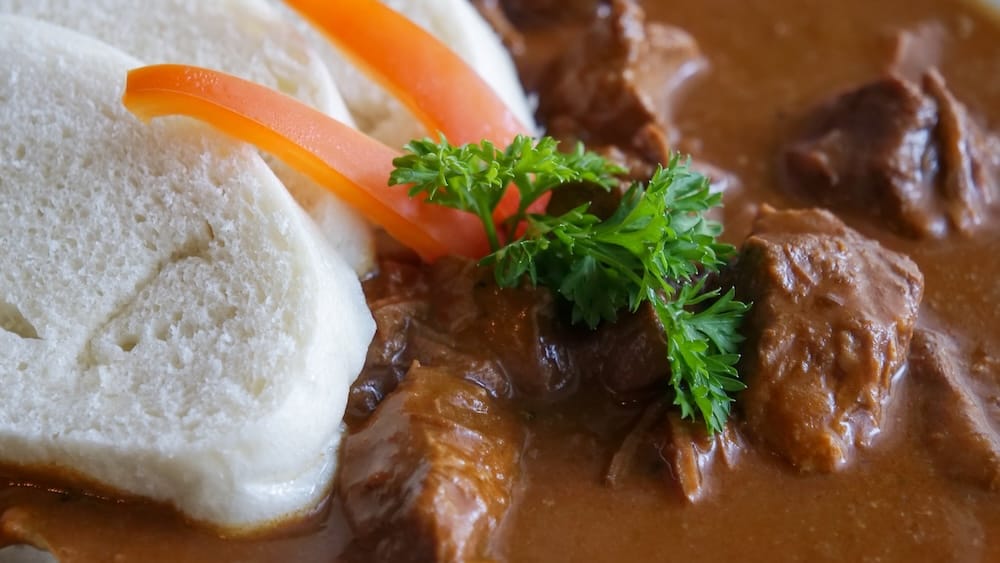 Primer plano del goulash de carne en Bistro Praha en Edmonton, mostrando trozos tiernos de carne en una salsa marrón espesa, decorado con perejil y tiras de morrón rojo, junto a rodajas de dumpling de pan.