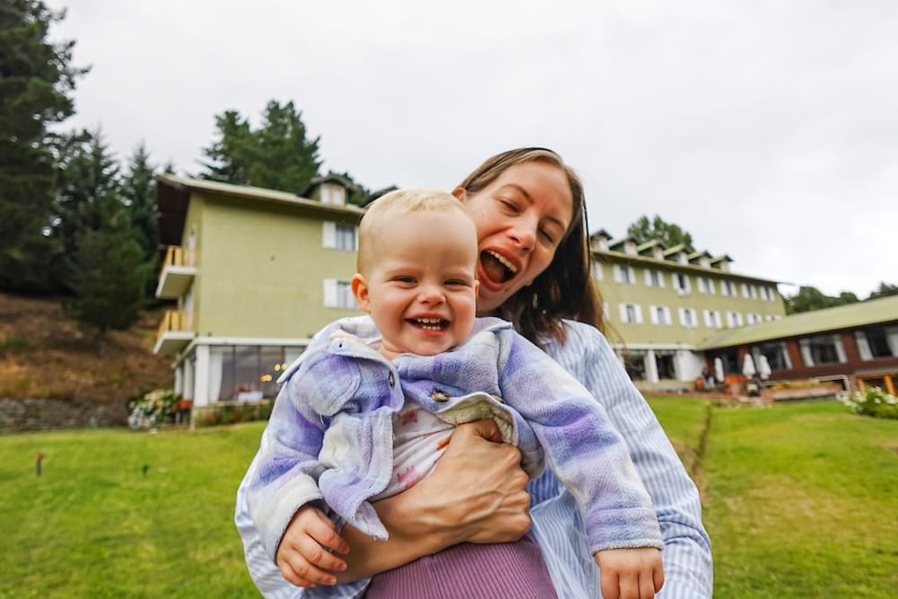 Audrey Bergner holds a smiling Aurelia Jeffery on the lawn outside Gran Hotel Panamericano in Bariloche, Río Negro, Patagonia, Argentina, with the green hotel building and forested grounds behind them.