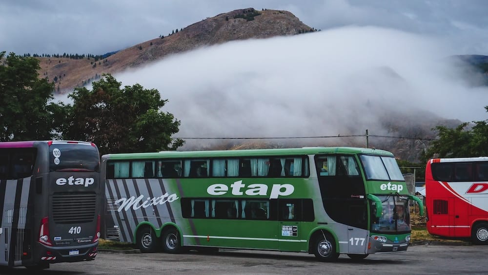 Green Etap coach bus parked in Esquel, Argentina, with other long-distance buses nearby and a low cloudbank hanging across the Andean hills in the background.