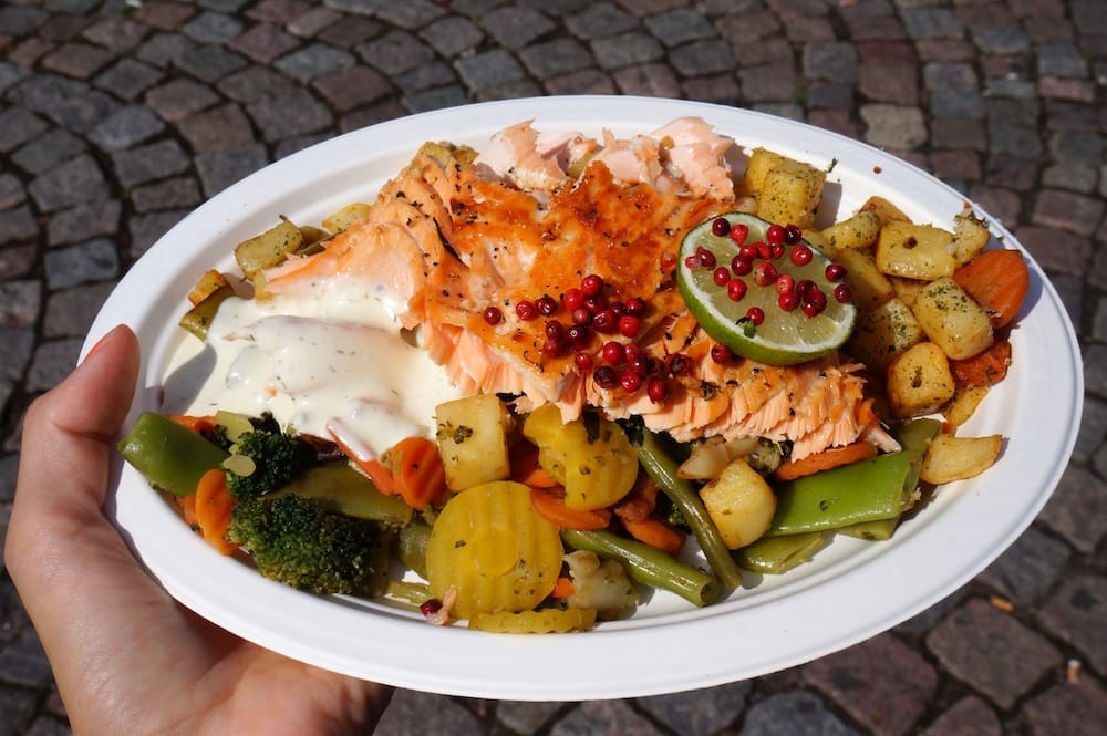 Grilled salmon served with roasted potatoes, mixed vegetables, garlic sauce, lime and red peppercorns at Helsinki Market Square, creating a colourful Finnish harbour lunch in summer.
