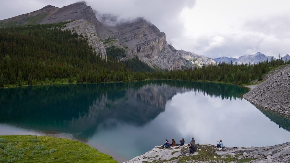 Pícnic en grupo, lago Matthias, Halfway Lodge, Parque Nacional Banff, Alberta, Canadá Un grupo pequeño se sienta en las rocas a hacer un pícnic junto al lago turquesa Matthias cerca de Halfway Lodge en el Parque Nacional Banff, Alberta, Canadá, con picos reflejados y nubes bajas.