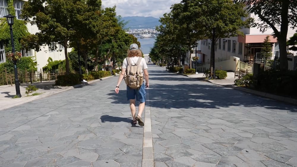 Samuel Jeffery walks down the broad stone-paved Hachimanzaka slope in Hakodate, Hokkaido, Japan, with trees and buildings lining the street and the harbour and distant mountains visible ahead.
