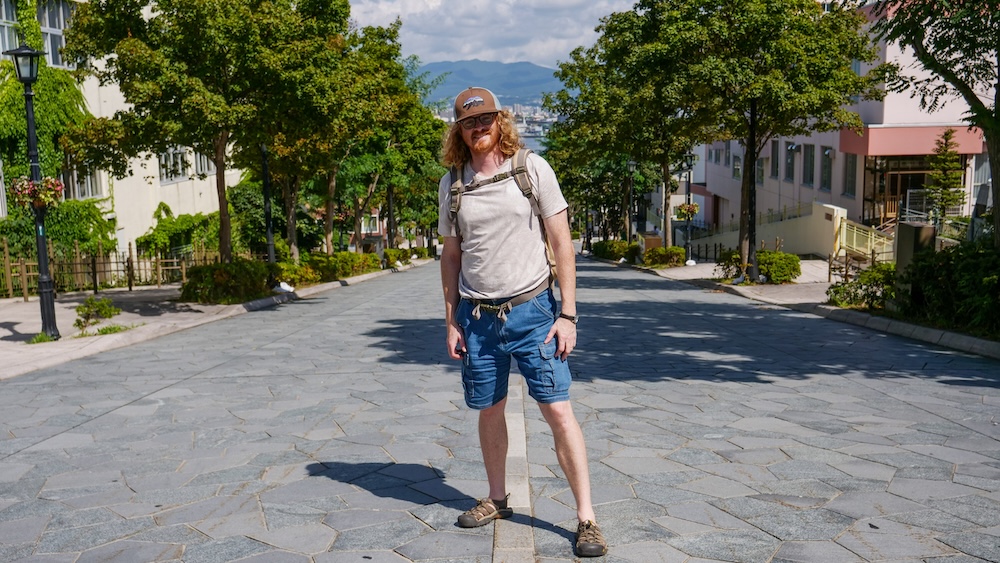 Samuel Jeffery on Hachimanzaka Slope in Hakodate, Hokkaido, Japan – Historic Street and Harbour Views Samuel Jeffery standing on the broad stone-paved Hachimanzaka slope in Hakodate, Hokkaido, Japan, wearing a cap, sunglasses and backpack, with leafy trees, buildings and a view toward the harbour behind him.