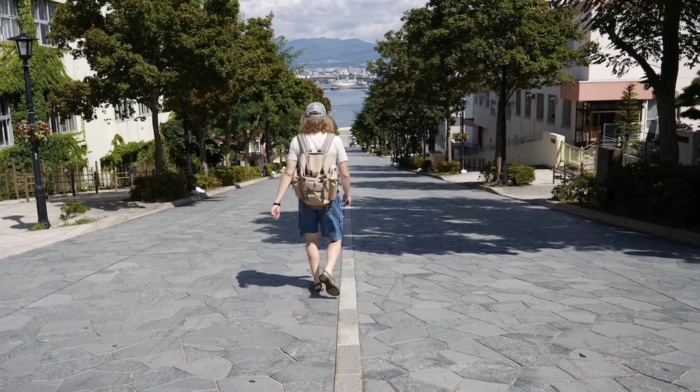 Samuel Jeffery Walking Down Hachimanzaka Slope Toward Hakodate Harbour in Motomachi, Hokkaido, Japan Samuel Jeffery wearing a cap and backpack walks downhill on the wide stone-paved Hachimanzaka Slope in Hakodate, Hokkaido, Japan, with leafy trees, buildings, the harbour and distant mountains visible ahead.