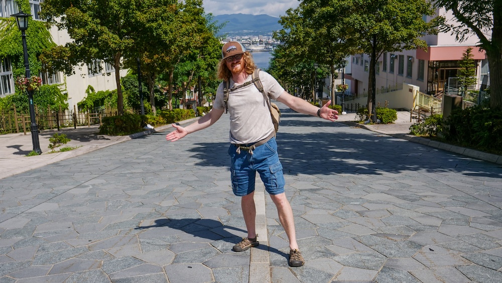 Samuel Jeffery standing on Hachimanzaka Slope in Motomachi, Hakodate, Hokkaido, Japan, with the stone-paved street descending toward Hakodate Port and the harbour visible below.