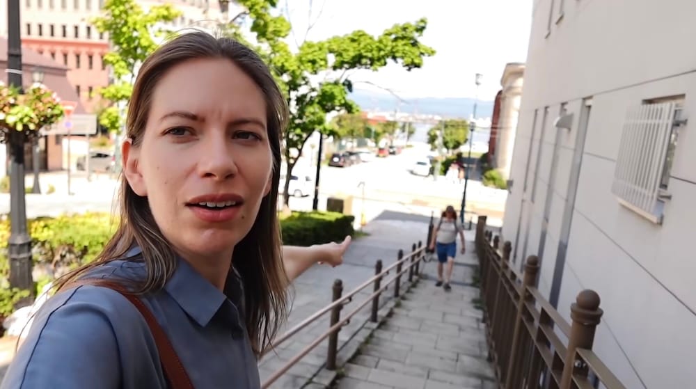 Stairs beside Hachimanzaka slope in Motomachi, Hakodate, Hokkaido, Japan – Audrey Bergner and Samuel Jeffery Audrey Bergner points toward Samuel Jeffery walking on the railed stairs beside Hachimanzaka in Motomachi, Hakodate, Hokkaido, Japan, with harbour views and city buildings visible down the slope.