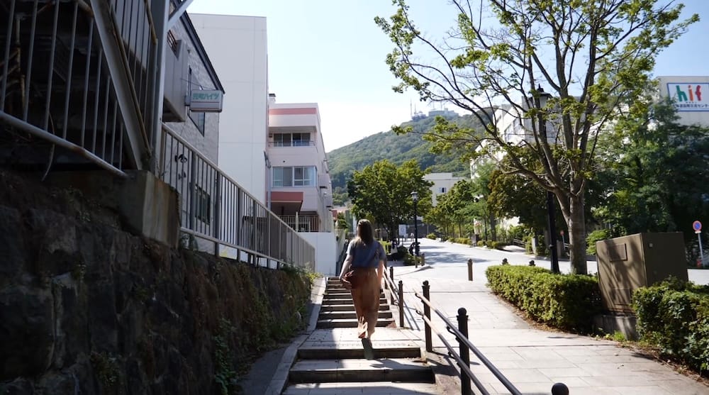 Climbing the stairs on Hachimanzaka, Hakodate, Hokkaido, Japan – Audrey Bergner Audrey Bergner climbs the stone stairs of Hachimanzaka in Motomachi, Hakodate, Hokkaido, Japan, with residential buildings, a tree-lined street and a hazy hill visible ahead.