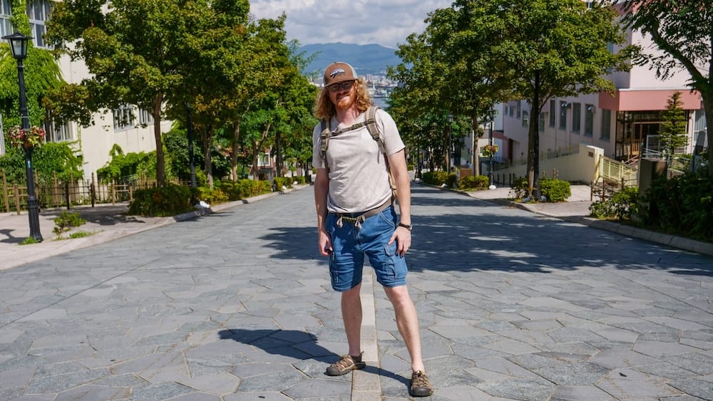 Hachimanzaka stone-paved slope in Motomachi, Hakodate, Hokkaido, Japan – Samuel Jeffery exploring on foot Samuel Jeffery stands on the wide stone-paved slope of Hachimanzaka in Hakodate, Hokkaido, Japan, wearing a backpack and cap, with tree-lined streets, buildings and hazy mountains visible in the distance.