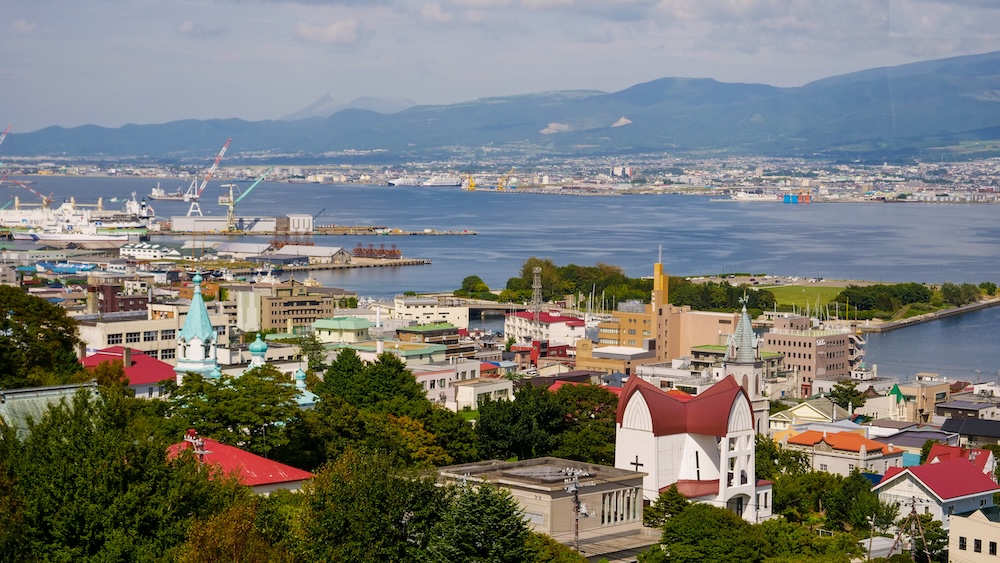 Hakodate Churches from the Ropeway, Hokkaido, Japan – Orthodox and Anglican Church Rooftops Above the Harbour Elevated view from the Hakodate Ropeway overlooking Motomachi in Hokkaido, Japan, with the green domes of Hakodate Orthodox Church, the red roof of St. John's Anglican Church, city buildings, harbour waters and distant mountains.