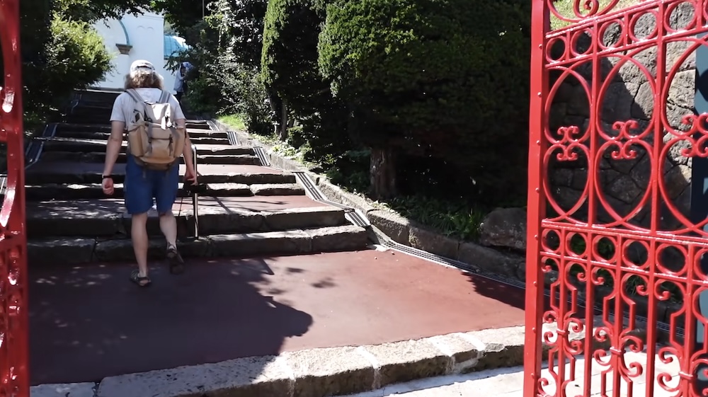 Samuel Jeffery Walking to Hakodate Churches in Motomachi, Hokkaido, Japan – Stone Steps and Red Entrance Gate Samuel Jeffery walking up a broad stone stairway through an ornate red iron gate in Motomachi, Hakodate, Hokkaido, Japan, carrying a backpack as he approaches the historic church area surrounded by trees and greenery.