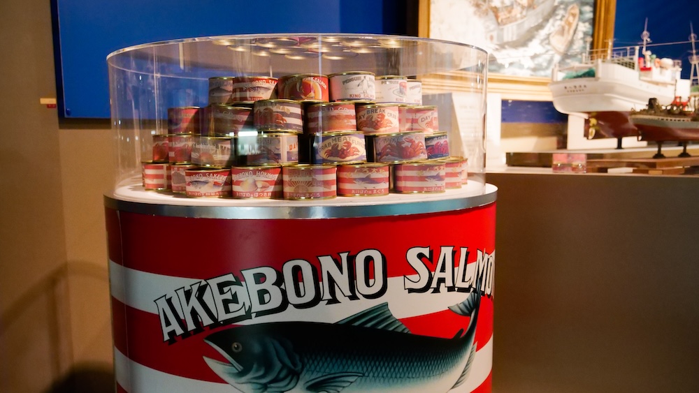Vintage Akebono salmon canned fish display, Hakodate City Hokuyo Museum, Goryokaku, Hokkaido, Japan Stack of vintage red-and-white striped canned fish under an acrylic dome on a large can-shaped pedestal reading Akebono Salmon, with ship models behind, at the Hakodate City Hokuyo Museum in Goryokaku, Hakodate, Hokkaido, Japan.