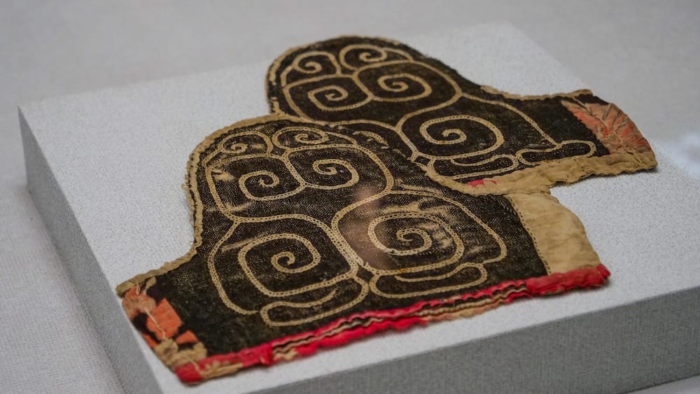 An embroidered textile with pale spiral patterns on dark cloth and red edging, displayed on a grey block at the Hakodate City Museum of Northern Peoples in Hakodate, Hokkaido, Japan.
