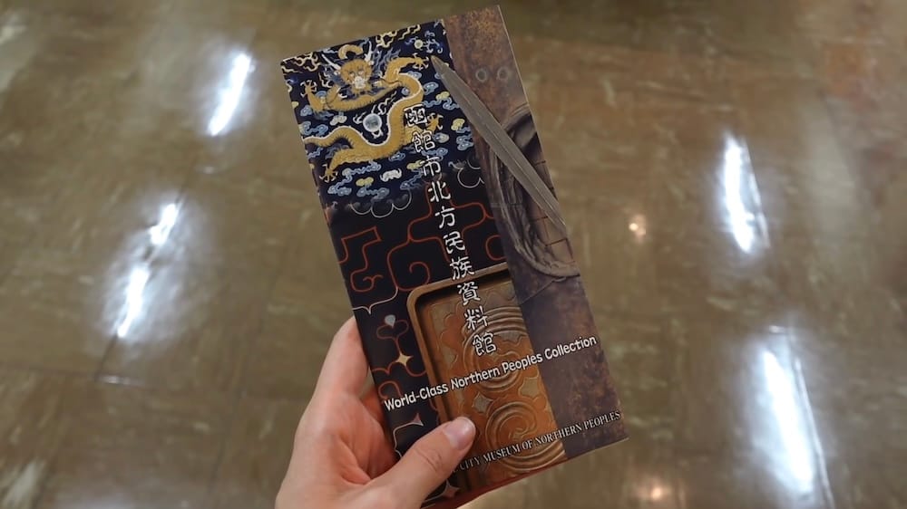 Audrey Bergner holds a Hakodate City Museum of Northern Peoples brochure inside the museum in Hakodate, Hokkaido, Japan, with the illustrated cover and polished indoor floor visible.