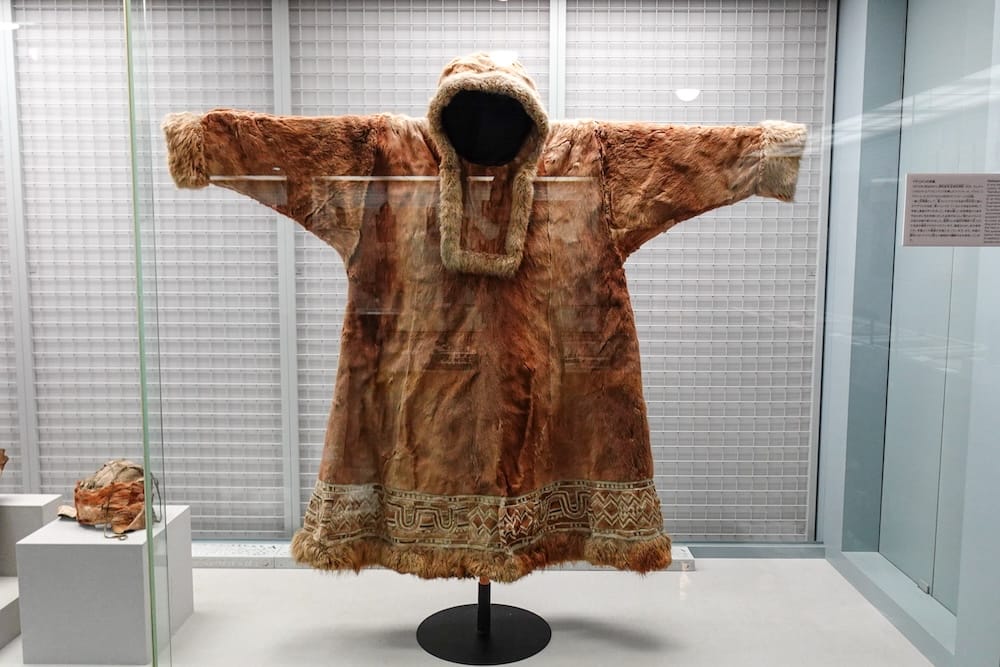 A fur-lined traditional garment is displayed behind glass inside the Hakodate City Museum of Northern Peoples in Hakodate, Hokkaido, Japan, with decorative trim visible around the hem and hood.